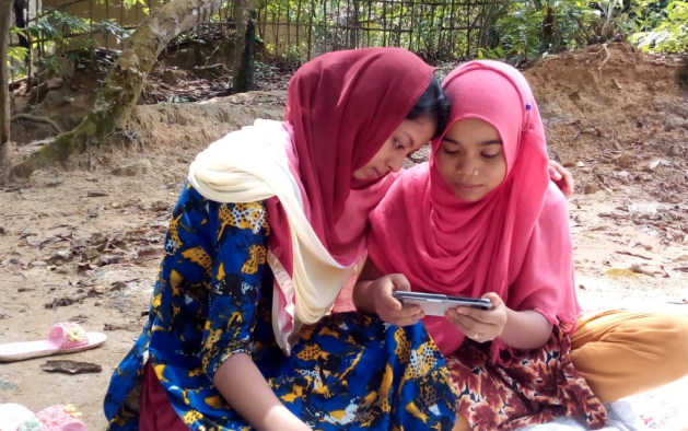 Two Rohingya girls sitting down on the floor next to each other. Their heads are very close together as they look at a the screen of a mobile phone that one of them is holding.
