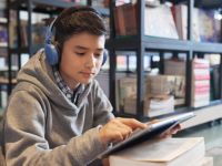 A young boy wearing headphones and holding a tablet in his hand. It looks like he's working on something in the tablet.