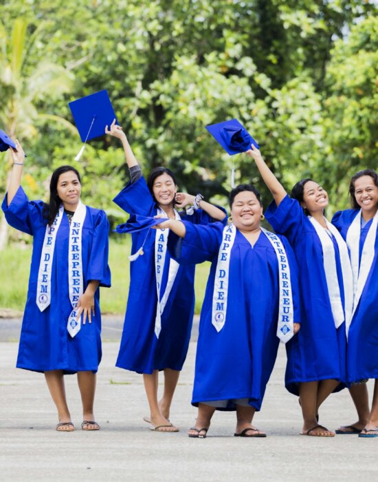 Five happy College of Micronesia students in dark blue graduation robes standing next to each other on a path, waving their mortarboards to celebrate graduating.