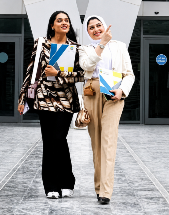 Two Kuwait University students walking outside the campus, smiling and holding books.