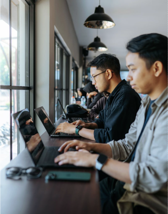 Alunos trabalhando em laptops em uma mesa comprida em frente a uma grande janela.